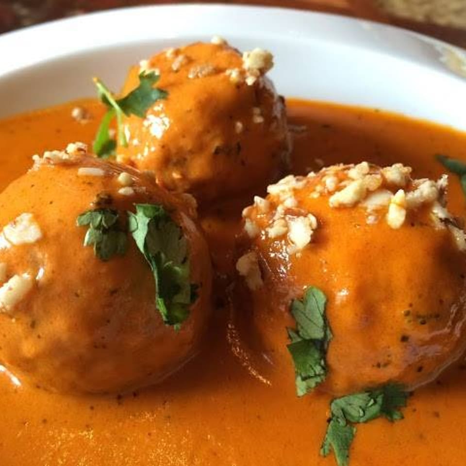 Three Indian Malai Kofta dumplings in a rich orange sauce, garnished with cilantro and nuts.
