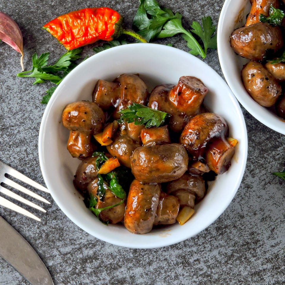 Two white bowls filled with Indian-style sautéed mushrooms, garnished with parsley, on a gray surface.