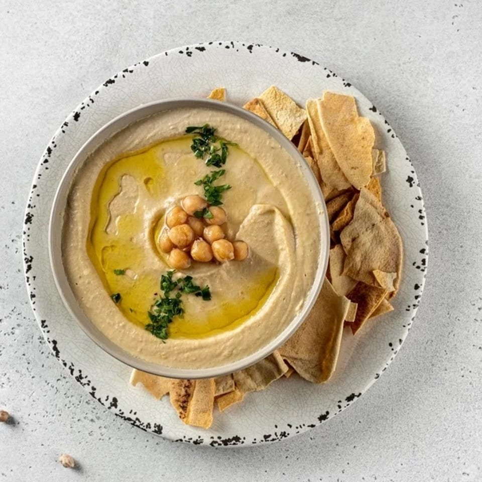 Creamy hummus topped with chickpeas, olive oil, and parsley, served with pita bread on a rustic plate.